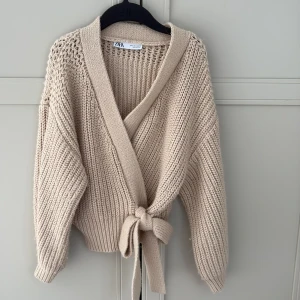 Beige stickad kofta från Zara - Säljer en mysig beige stickad kofta från Zara. Den har en omlott-design med knytband framtill och långa ärmar. Perfekt för kyliga dagar!🤎🤍pris kan diskuteras 