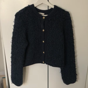 Mörkblå kofta med knappar - Säljer en mysig mörkblå kofta med guldknappar. Koftan har en fluffig textur och långa ärmar, perfekt för kyliga dagar. Den är både stilren och bekväm, ett måste i garderoben!