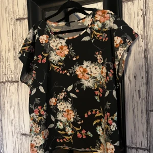 Blommig topp från Shein - Säljer en blommig topp från Shein i svart med färgglada blommor i orange, rosa och vitt. Toppen har korta ärmar och en rund halsringning. Perfekt för en färgglad look!