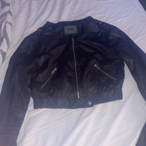 Svart jacka från ONLY - Snygg svart jacka från ONLY med dragkedja framtill och två snedställda fickor med dragkedjor. Jackan har en croppad design och är perfekt för en cool look. Den är tillverkad i ett glansigt syntetmaterial som ger en modern touch.