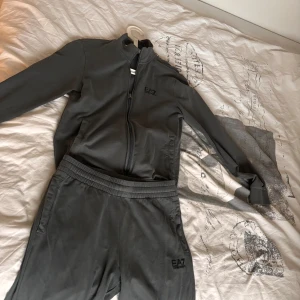 Grå träningsset från EA7 Emporio Armani - Säljer ett stilrent grått träningsset från EA7 Emporio Armani. Setet består av en tröja med dragkedja och matchande byxor. Perfekt för en sportig look eller avslappnad dag. Logotypen är diskret broderad på både trojan och byxorna.