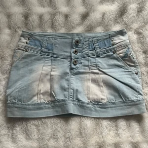 Ljusblå jeanskjol med knappar - Trendig ljusblå jeanskjol med knappar framtill och fickor både fram och bak. Kjolen har en kort längd och en cool tvättad look. Perfekt för en avslappnad stil. Midjemått 36cm och längd 30cm