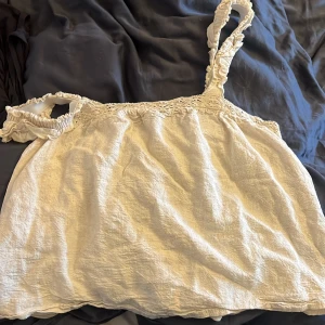 Vit ärmlös topp med spetsdetaljer - Säljer en söt vit ärmlös topp med spetsdetaljer vid axelbanden. Toppen har en luftig passform och är perfekt för varma dagar. Den är gjord i ett lätt och bekvämt material.