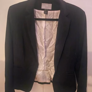 Svart kavaj från H&M - Snygg svart kavaj från H&M i storlek 40. Tillverkad av en blandning av polyester, ull och viskos med en bekväm passform. Perfekt för en stilren look.