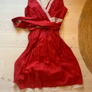 Röd sommarklänning med brodyrer - Säljer en röd omlottklänning med v-ringad hals och fina spetsdetaljer. Klänningen har en knytning i midjan och är ärmlös, perfekt för sommarens varma dagar. Jag är 167 och den går precis till knäna på mig 💖