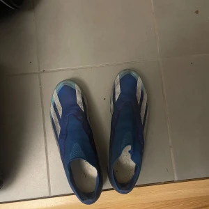 Blå fotbollsskor från Adidas - Säljer ett par blå fotbollsskor från Adidas med vita detaljer. Skorna har en strömlinjeformad design utan snörning för en tight passform. Perfekta för snabba rörelser på planen.