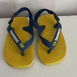Gula och blå flip-flops från Havaianas st.22 - Snygga flip-flops från Havaianas i gult och blått med brasiliansk flagga på remmen. Perfekta för sommardagar på stranden eller i stan. De har en bekväm passform och är lätta att bära.