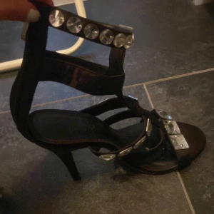 Svarta klackskor med strassdetaljer - Snygga svarta klackskor med remmar dekorerade med stora strassdetaljer. Perfekta för att ge en glittrig touch till din outfit. Skorna har en elegant klack och öppen tå. 