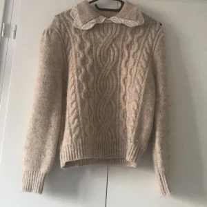 Beige stickad tröja med krage - Mysig beige stickad tröja med kabelstickat mönster och en söt krage. Perfekt för kyliga dagar och ger en klassisk look. Tröjan har långa ärmar och ribbade muddar vid ärmslut och nederkant.