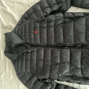 Svart pufferjacka från Polo Ralph Lauren - Säljer en snygg svart pufferjacka från Polo Ralph Lauren. Jackan har en dragkedja framtill och är långärmad. Den är quiltad för extra värme och har en liten röd logotyp på bröstet. Perfekt nu för våren 