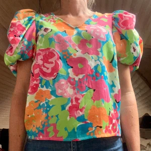 Färgglad blommig blus - Säljer en färgglad blus med blommigt mönster i rosa, grönt, blått och orange. Blusen har korta puffärmar och en v-ringad hals. Perfekt för att lysa upp garderoben!