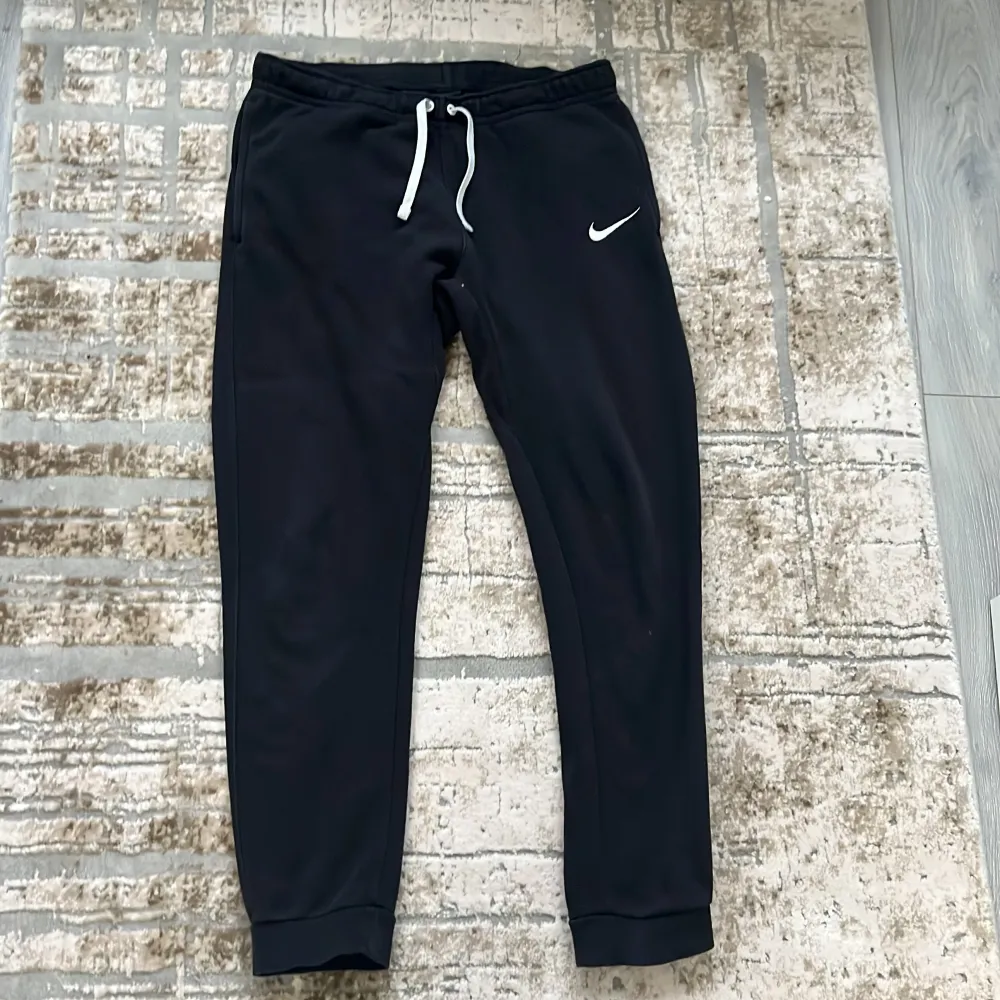 Säljer ett par svarta mjukisbyxor från Nike med vit dragsko i midjan och den klassiska Nike-loggan på benet. Byxorna har en bekväm passform och är perfekta för avslappnade dagar.. Farkut & Housut.