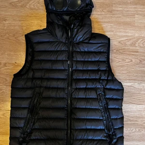 Svart dunväst från C.P. Company - Snygg svart dunväst från C.P. Company med en unik design. Västen har en dragkedja framtill och en huva med inbyggda glasögon. Perfekt för en stilren look. Strl 54