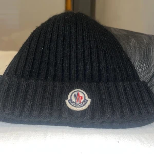Svart ribbad mössa från Moncler - Snygg svart ribbad mössa från Moncler med deras ikoniska logga framtill. Perfekt för att hålla värmen under kyliga dagar och ger en stilren look.