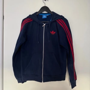 Mörkblå hoodie från Adidas - Säljer en mörkblå hoodie från Adidas med röda ränder på ärmarna och dragkedja framtill. Den har en justerbar huva med snören och den klassiska Adidas-loggan på bröstet. Perfekt för en sportig look!