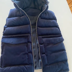 Mörkblå vendiera väst.  - Snygg mörkblå väst med quiltad design och dragkedja framtill. Västen har en huva och stora fickor med lock. Perfekt för att hålla värmen under kyliga dagar.