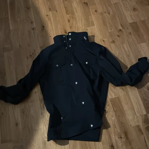 Mörkblå jacka från J.Lindeberg - Säljer en stilren mörkblå jacka från J.Lindeberg. Jackan har en hög krage och knappar framtill. Den är långärmad och har två bröstfickor med lock. Perfekt för en snygg och avslappnad look. Ny pris 3000kr