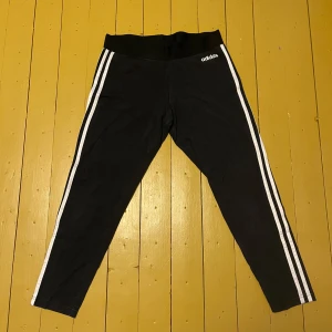 Svarta leggings från Adidas - Snygga svarta leggings från Adidas med klassiska vita ränder längs sidorna i stl L. Perfekta för träning eller en avslappnad dag. De har en elastisk midja för extra komfort. Använda ett fåtal gånger och är i bra skick 😊