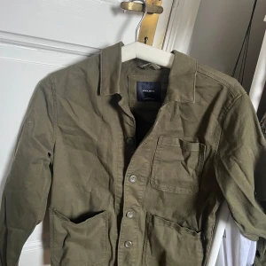 Olivgrön jacka från Jack & Jones - Säljer en olivgrön jacka från Jack & Jones med knappar framtill. Jackan har flera praktiska fickor och en klassisk krage. Perfekt för en avslappnad stil.