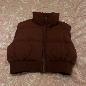 Brun dunväst från Zara - Säljer en brun dunväst med dragkedja. Västen har en hög krage och elastisk nederkant för en bekväm passform. Perfekt för kyligare dagar.