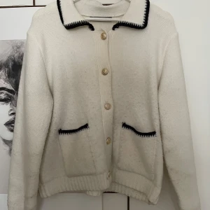 Vit kofta med svarta detaljer från BikBok - Vit kofta från BikBok med svarta detaljer på krage och fickor. Guldiga knappar framtill och två fickor. Klassisk krage och ribbade muddar. 