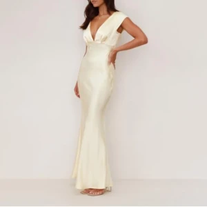 Champagnefärgad långklänning i satin - Aldrig använt då jag råka köpa två av samma. Elegant långklänning i champagnefärgad satin med djup v-ringning och öppen rygg. Klänningen har korta ärmar och går in lite vid midjan. Perfekt för bal eller festliga tillfällen. 