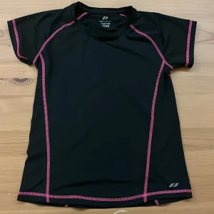 Svart tränings-tshirt Pro Touch - Sportig svart t-shirt från Pro Touch med rosa kontrastsömmar. Perfekt för träning, tillverkad i syntetmaterial som andas. Kortärmad modell med rund hals och reflekterande detaljer på ryggen. Sitter skönt och är lätt att röra sig i.