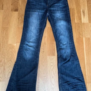 Blå bootcut jeans med slitna detaljer - Snygga blå jeans med bootcut passform och lätt slitna detaljer och justerbar midja. Jeansen har klassisk femficksdesign, normal midja och är tillverkade i denim. Perfekta för dig som gillar en retro vibe och vill ha ett par jeans med lite extra vidd nertill.