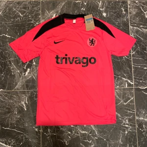 Chelsea tränings t-shirt Nike M - Chelsea FC tränings t-shirt från Nike i stark rosa med svarta detaljer på axlar och krage. Trivago-logga och klubbmärke på bröstet. Tillverkad i lätt och ventilerande polyester, perfekt för fotbollsträning eller match.