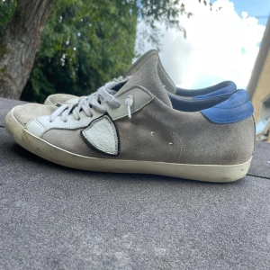 Grå och vita sneakers med blå detalj - Sneakers i mocka och skinn med grå och vita paneler samt blå häl. Klassisk snörning och rund tå. Snygg kontrast mellan material och färger, perfekt för en avslappnad streetstil. Storlek 42. Pris är 799 och inte mindre