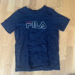 Svart FILA t-shirt  - Svart t-shirt från FILA med stor, vit och röd logga på bröstet. Klassisk passform med rund hals och korta ärmar. Tillverkad i mjuk bomull som är skön att ha på sig hela dagen. Perfekt för dig som gillar sportiga och stilrena plagg.