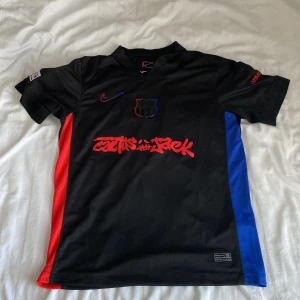 FC Barcelona x Cactus Jack svart matchtröja - Snygg svart FC Barcelona x Cactus Jack fotbollströja från Nike med röda och blå detaljer på sidorna. Tryck med Cactus Jack-logga och UNHCR på ryggen. Kortärmad modell i lätt och ventilerande material, perfekt för dig som vill sticka ut på planen.