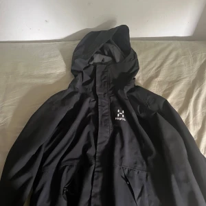 Svart vindjacka från Haglöfs, S - Svart vindjacka från Haglöfs med huva och dragkedja framtill. Jackan har diskret logga på bröstet och är tillverkad i återvunnen polyester. Perfekt för blåsiga dagar och enkel att matcha med allt. Snygg, funktionell och stilren design.
