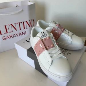 Valentino Rockstud sneakers vit/rosa - Snygga Valentino Rockstud sneakers i vitt skinn med bred rosa rem över vristen dekorerad med ikoniska nitar. Klassisk rund tå, platt sula och vita snören. Perfekta för dig som vill ha en lyxig och trendig touch till din outfit.
