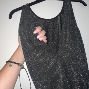 Svart glittrig kortklänning - Supersnygg svart kortklänning med glittrigt tyg som verkligen fångar ljuset. Ärmlös modell med rund halsringning och markerad midja. Perfekt för dig som vill sticka ut med en festlig look. Materialet känns mjukt och stretchigt. Nyskick. Kan användas åt båda sidor med antingen djup urringning eller inte❤️
