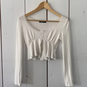 Vit croppad bolerotopp från Gina Tricot - Supersöt vit croppad topp från Gina Tricot med långa ärmar, spetsdetaljer längs kanterna och liten rosett framtill. Toppen har en volangig nederdel. Perfekt att styla med jeans eller kjol.