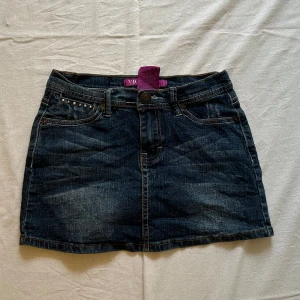 Mörk jeanskjol från Vigoss med nitar - Midjemått rakt över: 31 cm, längd: 29 cm Snygg mörkblå jeanskjol från Vigoss med coola nitar på fickorna och broderade detaljer bak. Klassisk femficksmodell med bälteshällor och slitningar för en trendig look. Perfekt till sneakers eller boots för en chill stil.