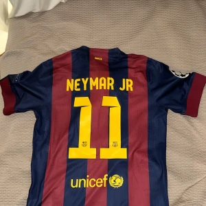 FC Barcelona Neymar Jr 11 matchtröja - Säljer en officiell FC Barcelona matchtröja från Nike med Neymar Jr och nummer 11 på ryggen. Tröjan är randig i mörkblått och rött, har korta ärmar och är tillverkad i lätt, ventilerande polyester. Framsidan har Qatar Airways-logga och klubbmärke, baksidan har Unicef-tryck.