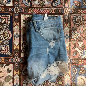 Jeansshorts med spets och slitningar - Snygga blå jeansshorts med slitna detaljer och råa kanter. Nedtill finns vit spets som sticker ut. Ändats använda ett fåtal gånger, jättebra skick.