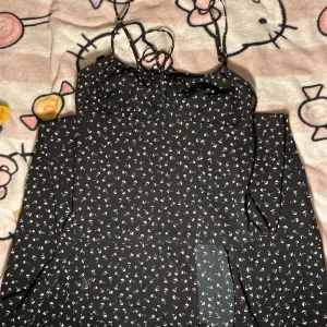 Svart blommig långklänning med slits - Supersöt svart långklänning med små vita blommor, smala axelband och en snygg slits framtill. Klänningen har en liten knytning vid bysten och är perfekt för dig som gillar en feminin och trendig look. Materialet känns lätt och flowy.