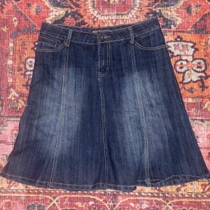 Jeanskjol med broderade fickor - Snygg jeanskjol från Tanna i mörkblå denim med utsvängd form, går typ till knäna. Kjolen har fickor fram och bak,  bakfickorna är dekorerade med coolt broderi. Storlek XL men liten i storleken, passar mig som är M.