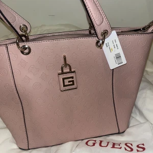 Guess rosa toteväska  - Säljer en snygg rosa toteväska från Guess, modell Kamryn. Väskan har präglat logomönster, guldfärgade metalldetaljer och dragkedja upptill. Insidan är fodrad med Guess-tryck och har flera fack, inklusive ett med dragkedja. Perfekt storlek för allt du behöver ha med dig.