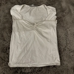 Vit topp - Söt vit topp med v-ringning och spetskant längs urringningen. Toppen har en rynkad detalj framtill som ger en feminin touch. Ärmlös modell i mjukt material, perfekt att styla med jeans eller kjol. Från lager 157 i storlek M