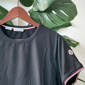 Moncler t-shirt  - Svart t-shirt från Moncler med rund hals och korta ärmar. Ärmsluten och halsringningen har snygga rödvita detaljer. Klassisk Moncler-logga på ärmen. Perfekt för dig som gillar stilrena och exklusiva plagg.