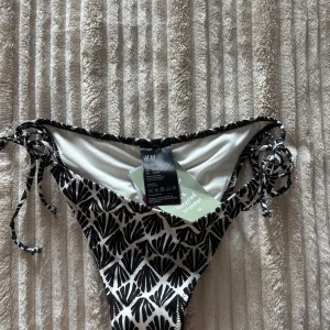 Svartvit bikiniunderdel från H&M - Snygg bikiniunderdel från H&M i svartvitt grafiskt mönster. Modellen har högt skuren midja och knytband i sidorna för perfekt passform. Tillverkad i återvunnen polyester, perfekt för strandhäng och pooldagar.