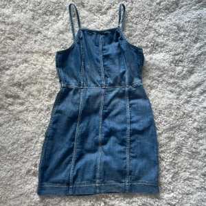 Jeansklänning med smala axelband H&M - Snygg jeansklänning från H&M med smala axelband och figurnära passform. Klänningen är i klassisk blå denim med synliga sömmar som ger en cool look. Perfekt att styla med sneakers eller boots för en trendig outfit.