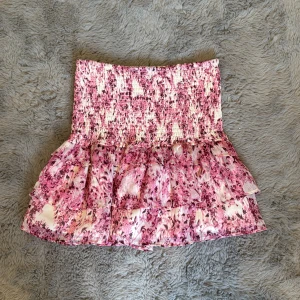 Rosa blommig volangkjol Gina Tricot - Supersöt kort kjol från Gina Tricot i rosa och vitt med blommigt mönster. Kjolen har smock i midjan och flera lager volanger som ger en fluffig och lekfull look. 
