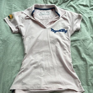 Ljusrosa pikétröja från Berkeley - Snygg ljusrosa pikétröja från Berkeley med blå broderad text på bröstet och coolt patch på ärmen. Klassisk krage, korta ärmar och figurnära passform. Står M men passar mer som en S, Perfekt för dig som gillar sportig och fräsch stil.