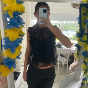 Svart spetsblus med volangdetaljer - Säljer en svart ärmlös blus i spets med volangdetaljer vid axlar och nederkant. Blusen har transparent spets över hela och en snygg hög halsringning. Perfekt för dig som vill ha en cool och feminin look.