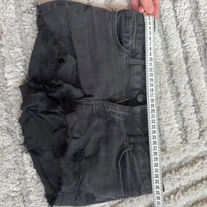 Svarta jeansshorts med fransig kant - Säljer ett par svarta jeansshorts  med fransig och sliten kant. Shortsen har klassisk femficksdesign, dragkedja och knapp framtill samt bälteshällor. Perfekta för sommarens alla äventyr och har en cool, avslappnad vibe.